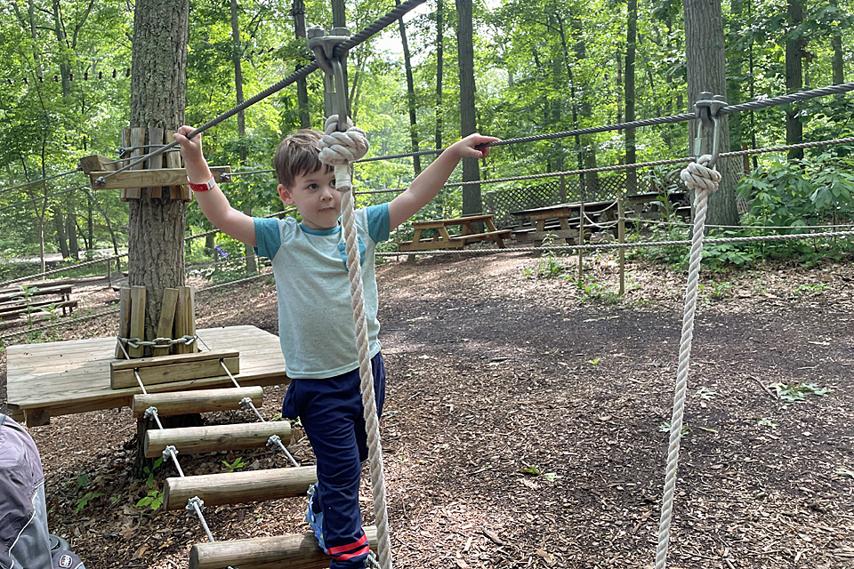Little boy crosses rope bridge at The Adventure Playground