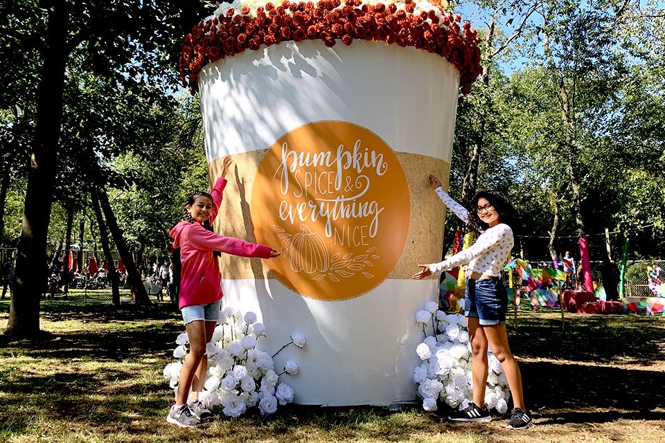 Two girls pose beside a larger-than-life pumpkin spice cup crafted out of flowers at The Fall Escape